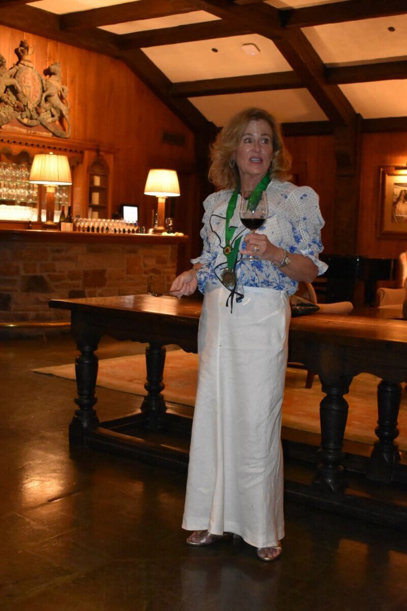 A woman in a long skirt and patterned blouse stands indoors holding a wine glass and eyeglasses, with a wooden table, warm lighting, and bar in the background.