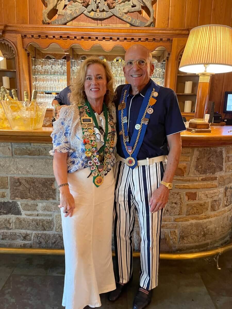 Pamela Hauber and a companion stand side by side in front of the bar, both wearing decorative chains and smiling at the camera. The bar has a stone base, wood paneling, and a lit lamp, creating a warm ambiance that highlights their happy faces.
