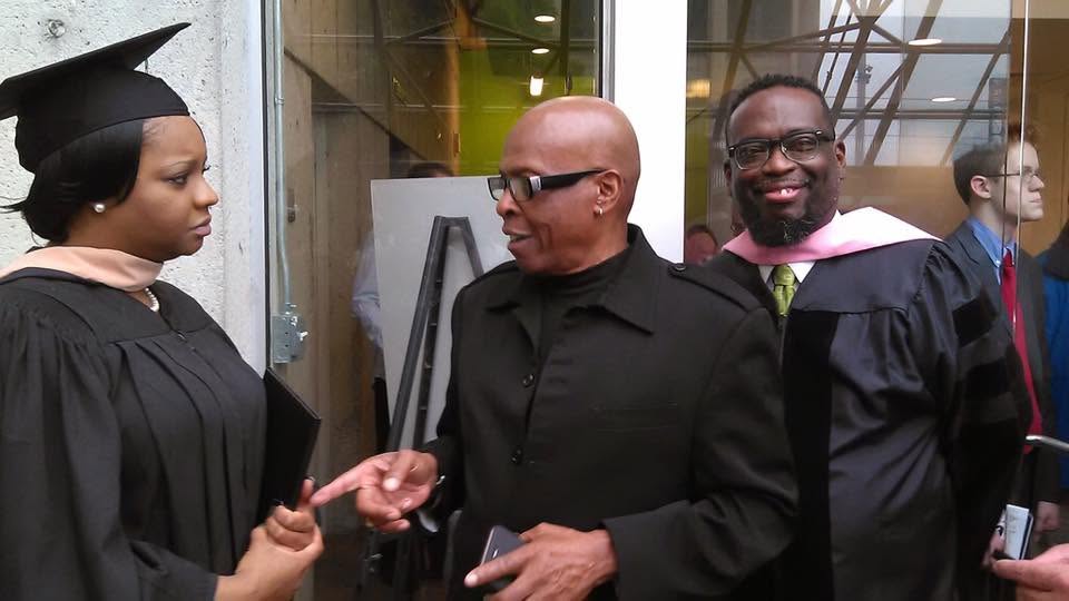 Two people in graduation gowns stand with a man in black clothing outdoors; one graduate appears to be receiving advice while another graduate smiles for the camera.