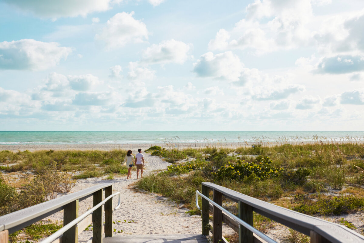 Beyond the Beach: A Weekend Exploring Fort Myers, Sanibel & Captiva - 2 Two people walk on a sandy path toward the ocean on Sanibel, surrounded by beach grass under a partly cloudy sky.