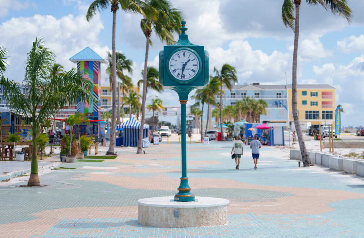 Beyond the Beach: A Weekend Exploring Fort Myers, Sanibel & Captiva - 7 Outdoor clock on a promenade in Fort Myers with palm trees, colorful buildings, and two people walking away in the distance under a partly cloudy sky.