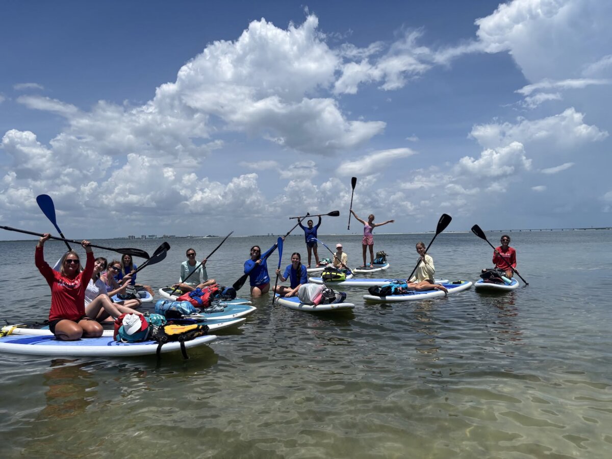 Beyond the Beach: A Weekend Exploring Fort Myers, Sanibel & Captiva - 4 A group of people sits and stands on paddleboards in shallow water near Captiva, holding up paddles, with partly cloudy skies and the distant land of Sanibel visible in the background.
