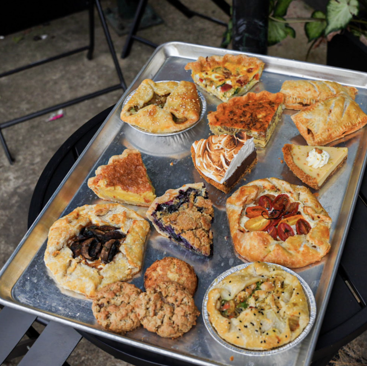 A metal tray on a black table holds an assortment of baked goods, including pies, tarts, cookies, and savory pastries, arranged in rows outdoors.