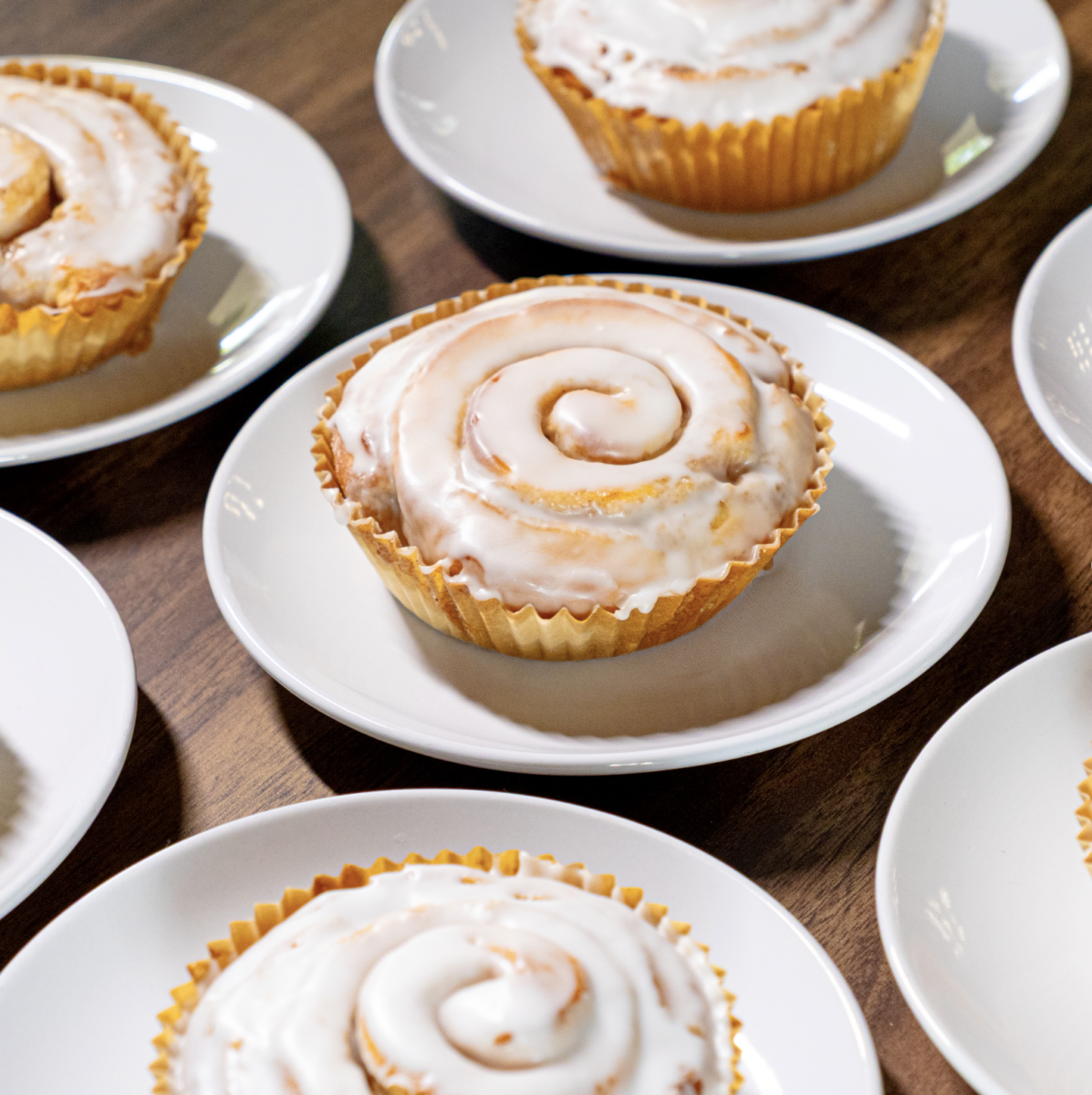 Iced cinnamon rolls sit on white plates, arranged on a wooden surface.