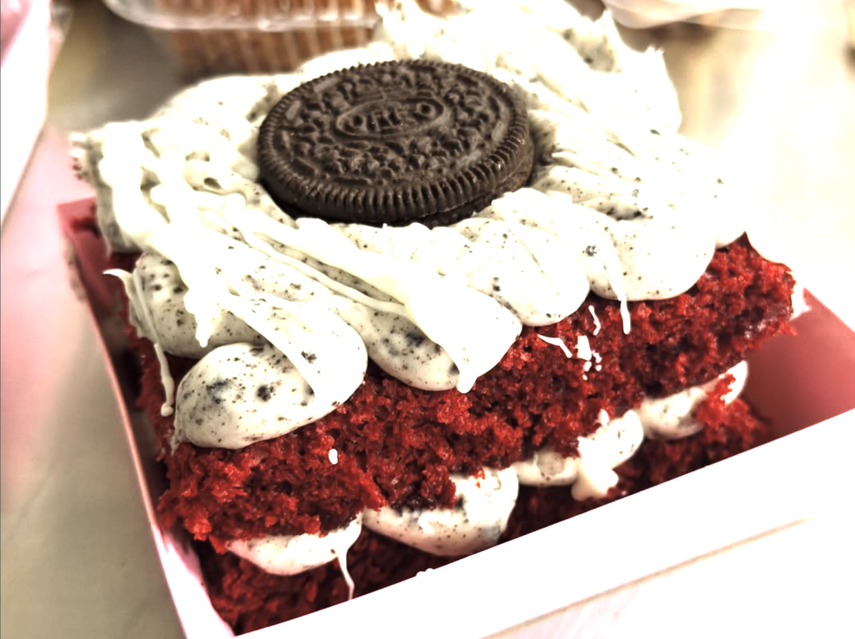 A slice of red velvet cake topped with cookies and cream frosting and an Oreo, served in a pink container.