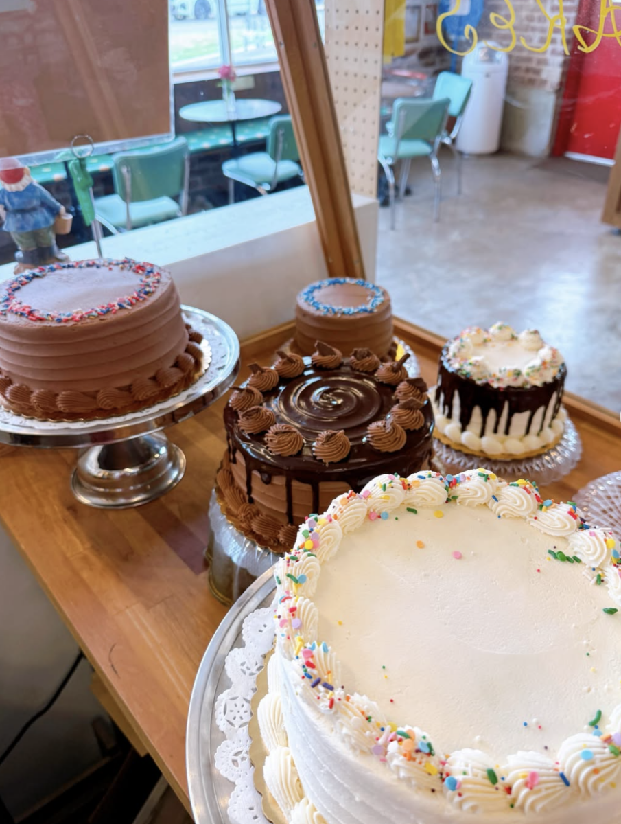 Four decorated cakes, including chocolate and vanilla varieties, are displayed on a wooden counter in a bakery with tables and chairs visible in the background.