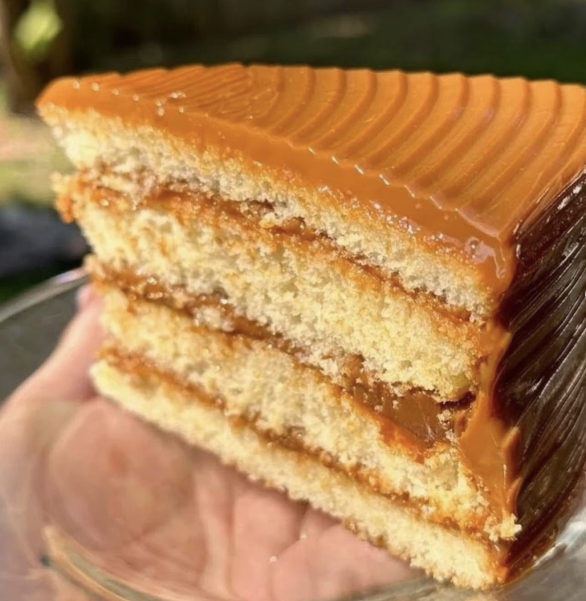 A close-up of a hand holding a slice of three-layer cake with caramel frosting and filling between each layer.