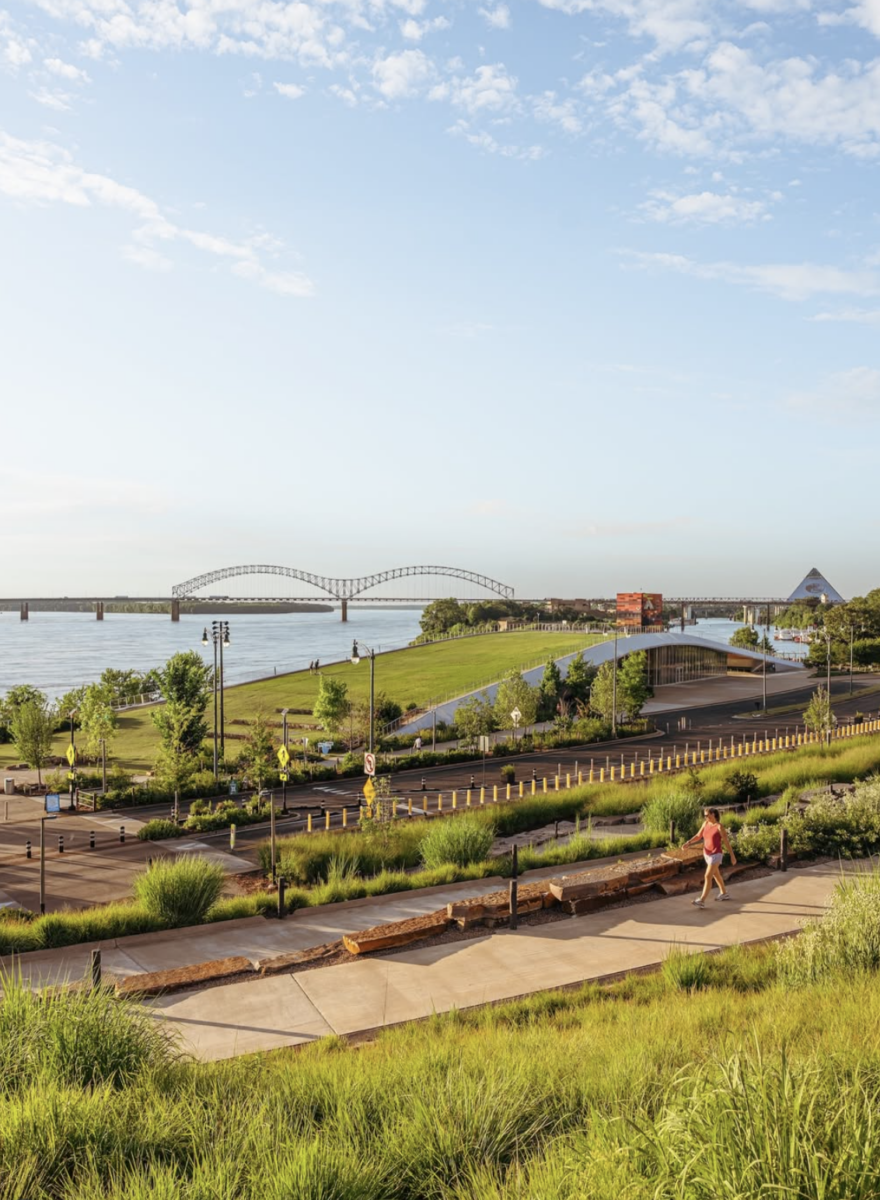 A person jogs along a riverside path with a bridge, river, and distant pyramid-shaped building visible under a blue sky.