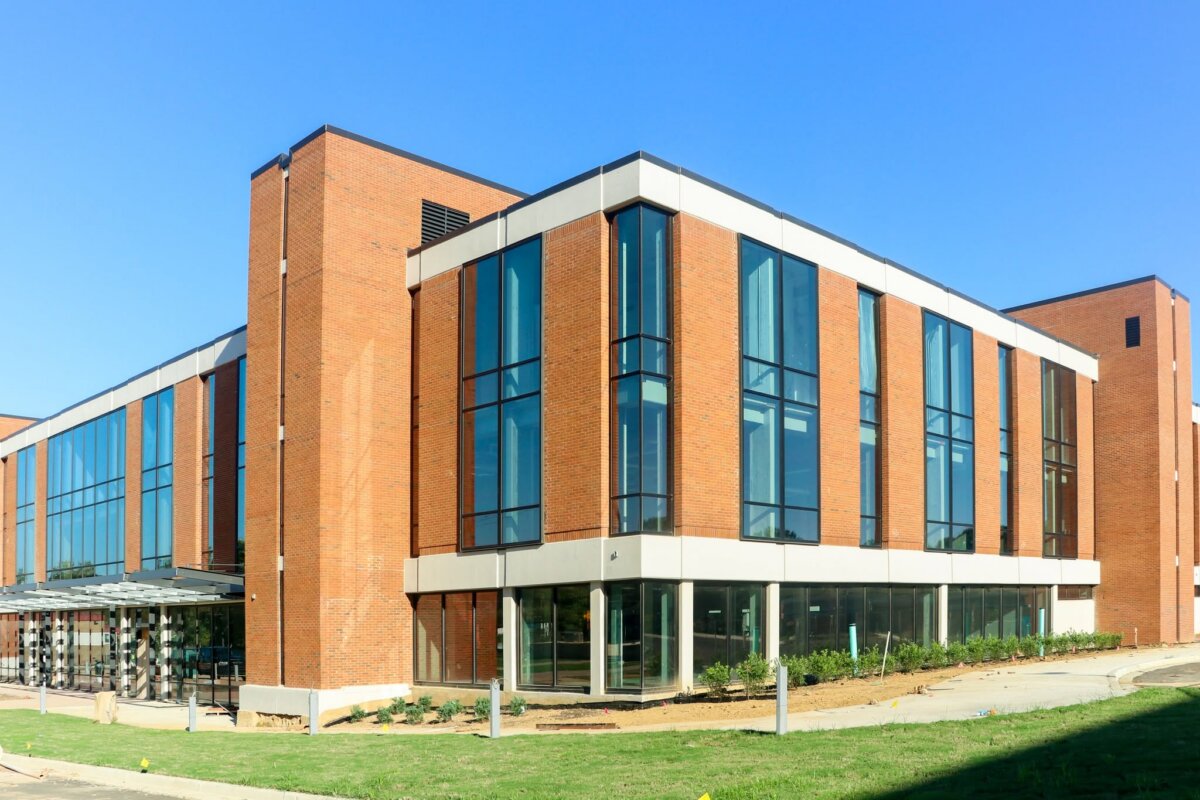 A modern, multi-story brick building with large glass windows, surrounded by grass and a clear blue sky.