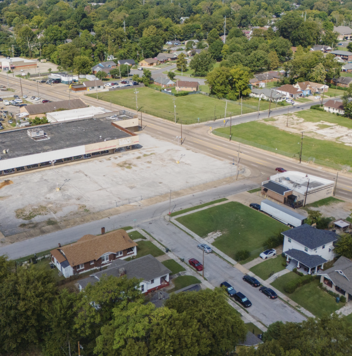 Aerial view of a suburban neighborhood with houses, a large empty lot, parking areas, streets, and trees.