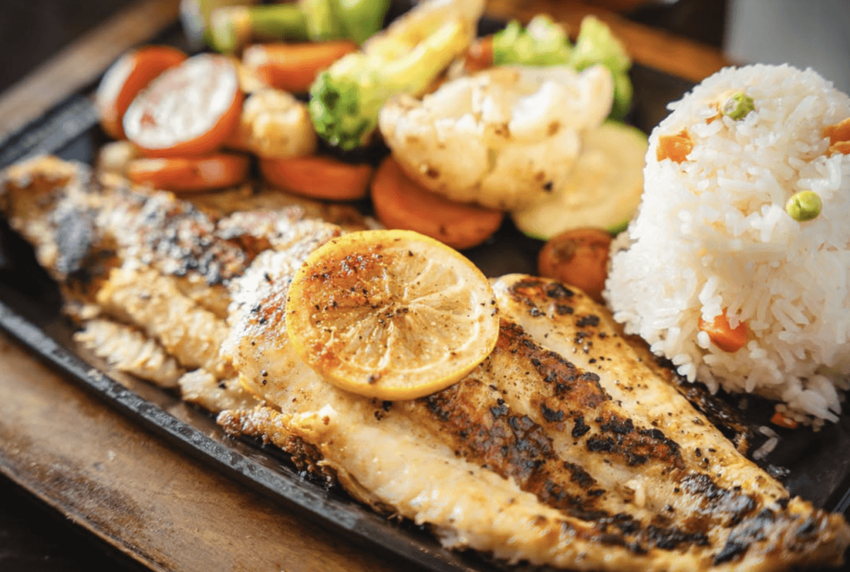Grilled fish fillet topped with a lemon slice, served with steamed rice and mixed vegetables including carrots, broccoli, zucchini, and cauliflower.