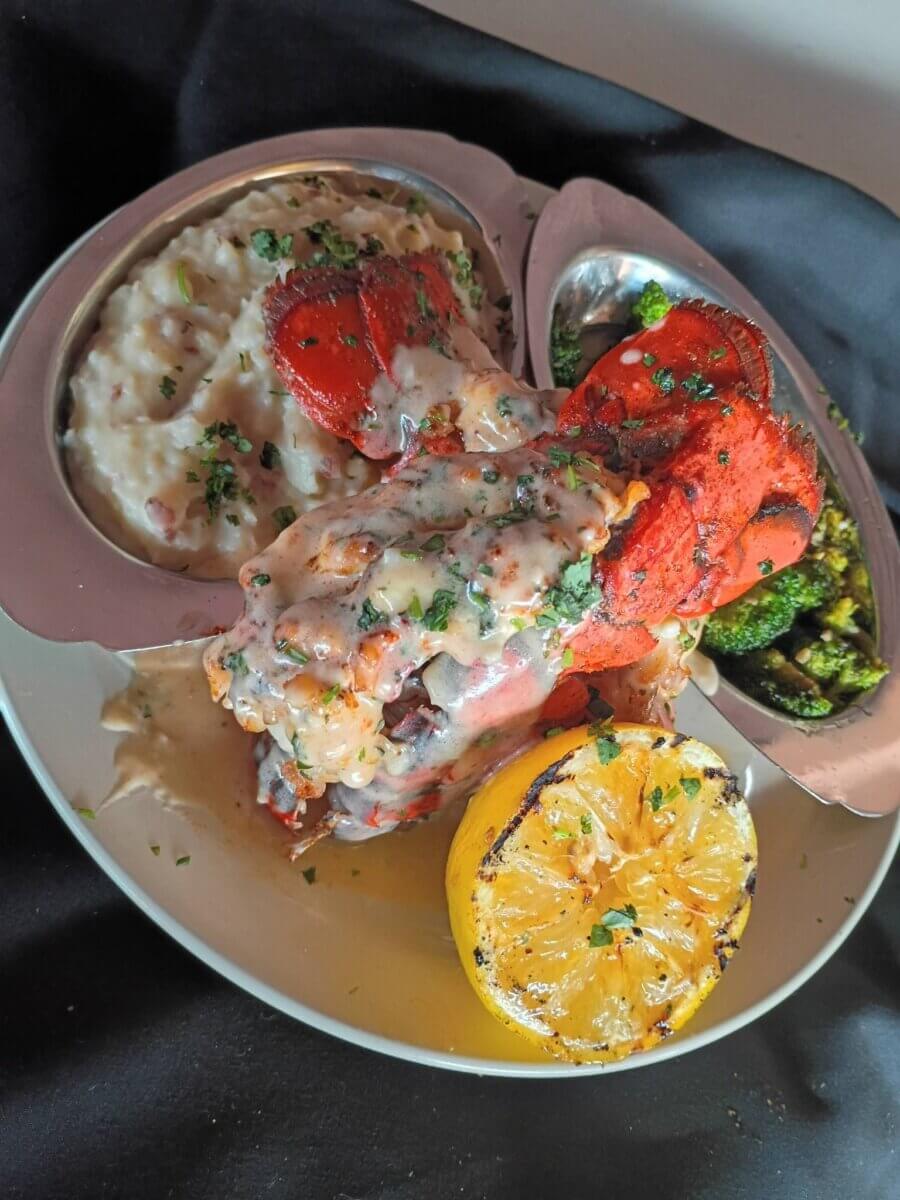 Lobster tail with creamy sauce, mashed potatoes, broccoli, and a grilled lemon served on a white plate with garnish.