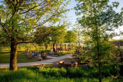 People gather at a park pavilion surrounded by green trees and grass on a sunny day, with picnic tables and winding walkways visible.