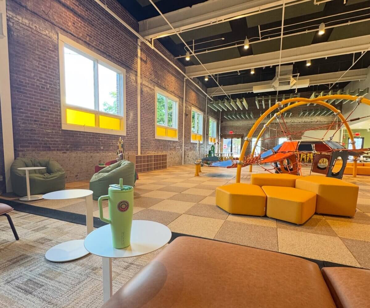 A modern indoor play area with climbing structures, yellow seating, bean bags, large windows, and a green tumbler on a small white table in the foreground—perfect for families exploring new Memphis restaurants or looking for unique spots among restaurants in Memphis.