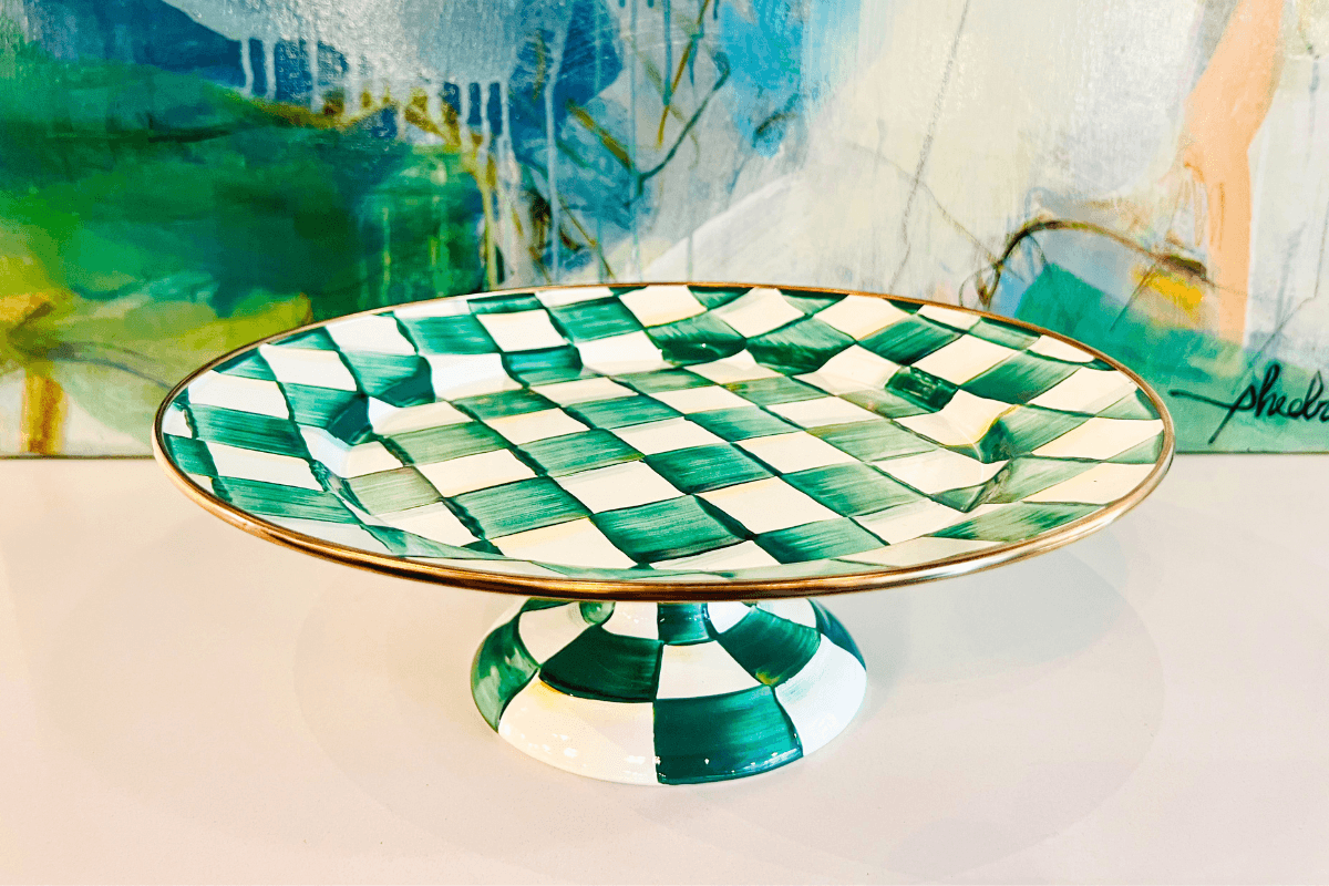 Green and white checkered ceramic cake stand with gold rim displayed on a white surface, colorful abstract painting in the background.