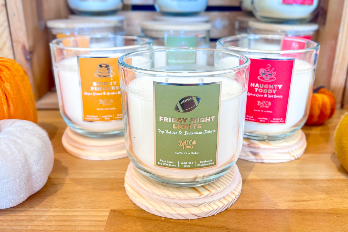 Three labeled candles in glass jars are displayed on wooden coasters, surrounded by decorative pumpkins on a wooden surface.