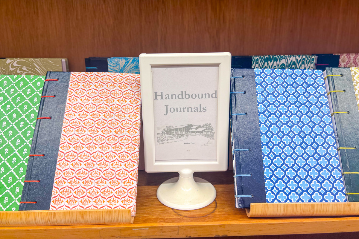 A display of handbound journals with patterned covers, placed on a wooden shelf, separated by a white sign reading “Handbound Journals.”.