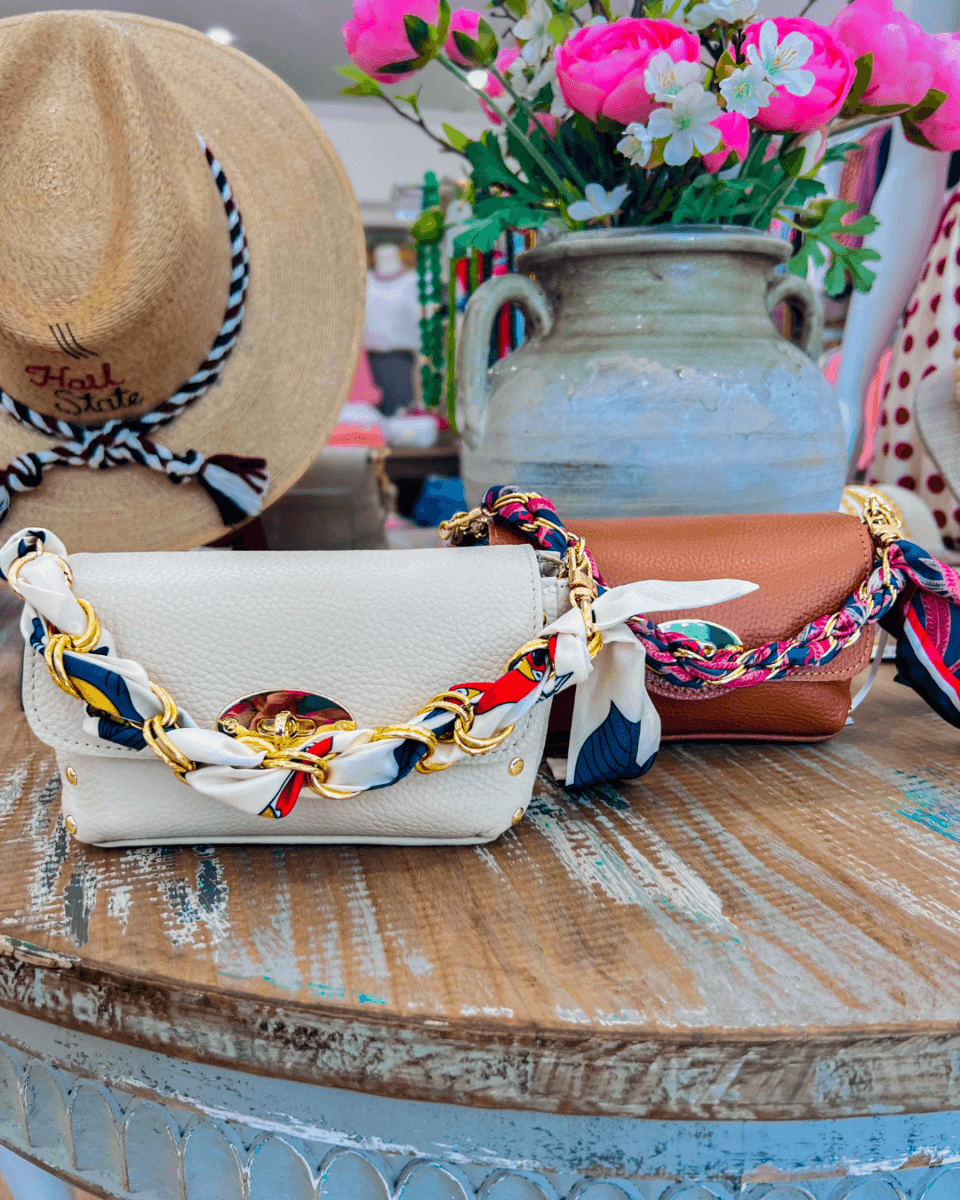 Two small handbags with colorful chain and scarf straps sit on a rustic round table beside a beige hat and a ceramic vase filled with pink flowers.