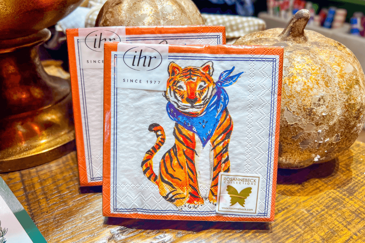 Two decorative napkins featuring an illustrated tiger with a blue bandana are displayed upright on a wooden surface near gold pumpkins.