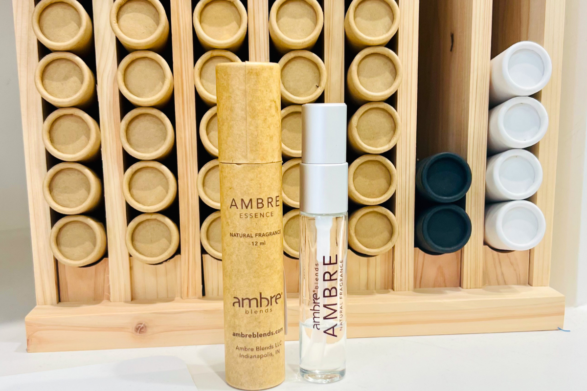 A cardboard tube and a glass spray bottle labeled “Ambre Essence” are placed in front of a wooden rack holding multiple cardboard and plastic tubes.