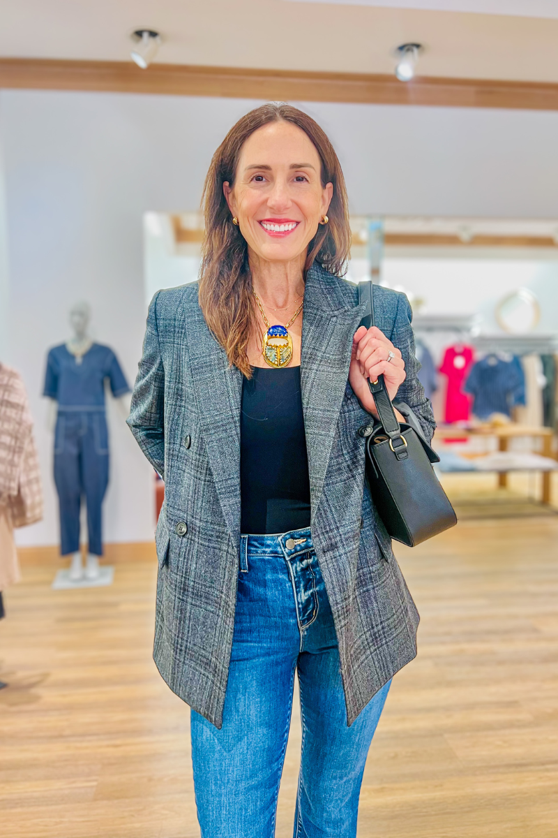 Woman stands indoors wearing a gray plaid blazer, black shirt, blue jeans, and holding a black handbag. Clothing racks and mannequins are visible in the background.