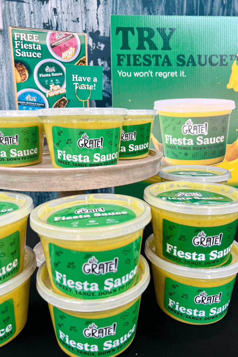 Plastic tubs of Grate! Fiesta Sauce are stacked on display with promotional signs offering free samples and encouraging people to try the sweet, tangy, down-to-earth sauce.