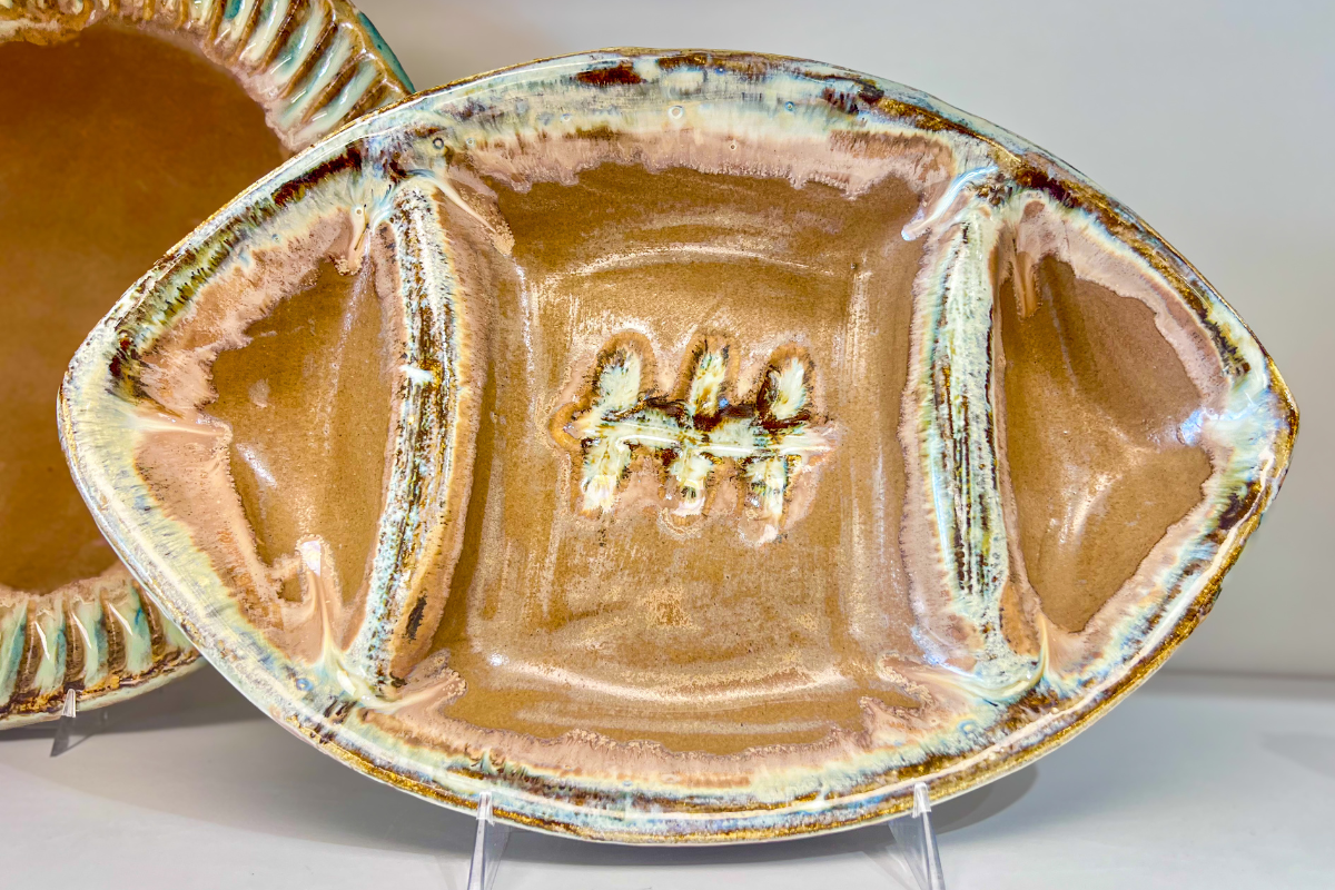 A ceramic platter shaped like a football with three vertical textured lines in the center, featuring brown and cream glaze, displayed on a stand.