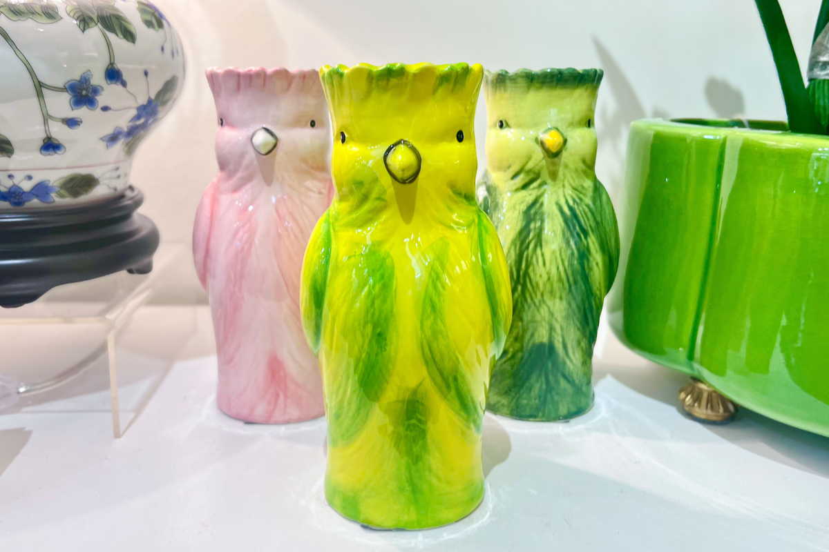 Three ceramic bird-shaped vases in pink, yellow, and green are displayed on a white surface among other ceramic items.