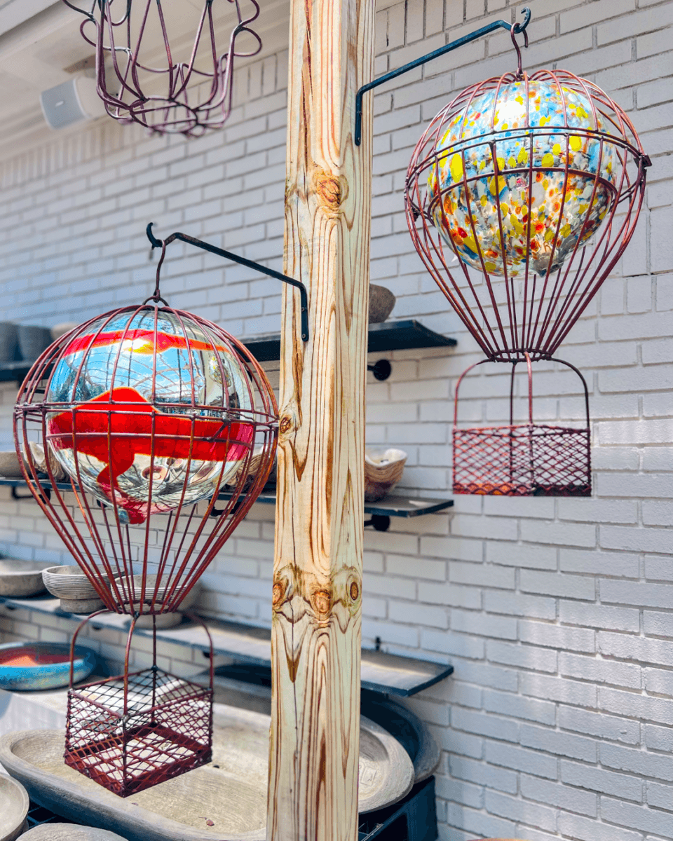 Two metal hot air balloon ornaments with mirrored and floral-patterned spheres hang on hooks attached to a wooden post in front of a white brick wall.