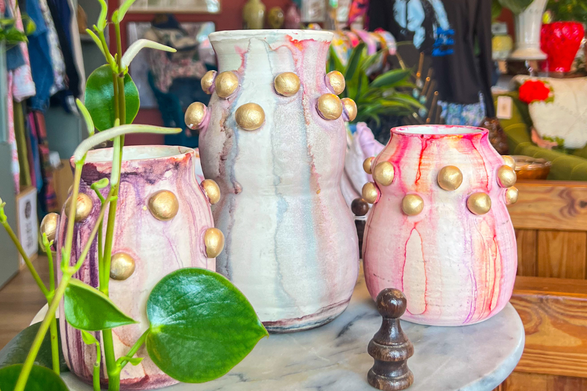 Three handmade ceramic vases with gold stud accents and pink marbling displayed on a round marble surface amid green plants in a shop setting.