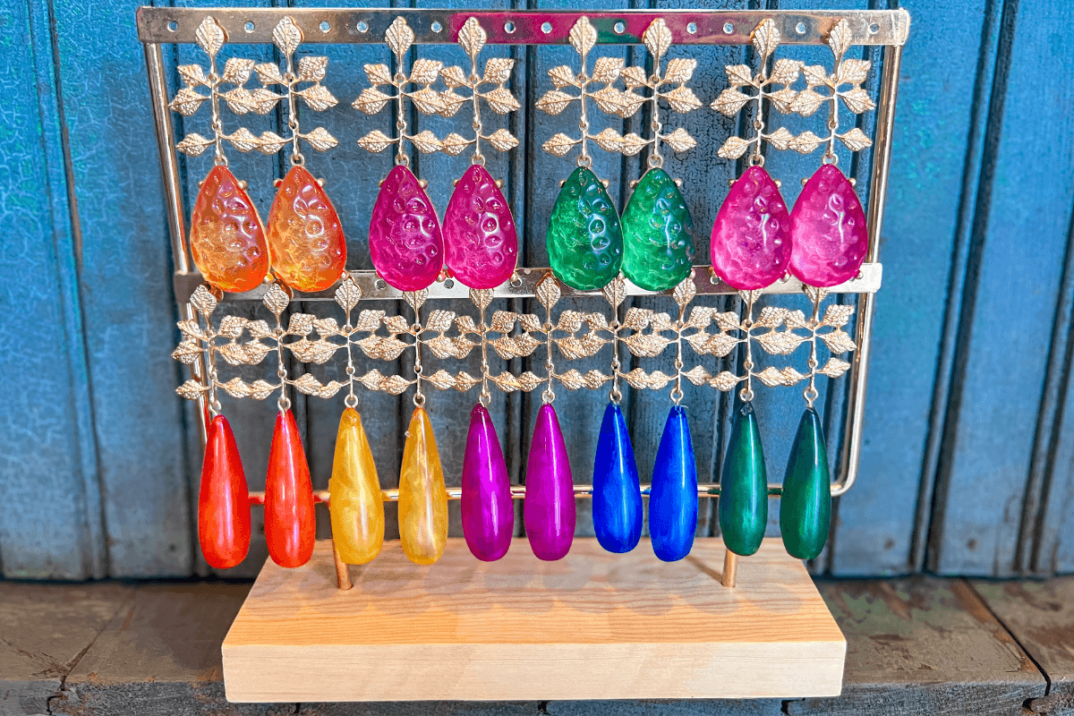 Colorful teardrop-shaped glass ornaments in orange, red, green, pink, yellow, purple, blue, and green, displayed on a decorative gold and wooden stand against a blue background.