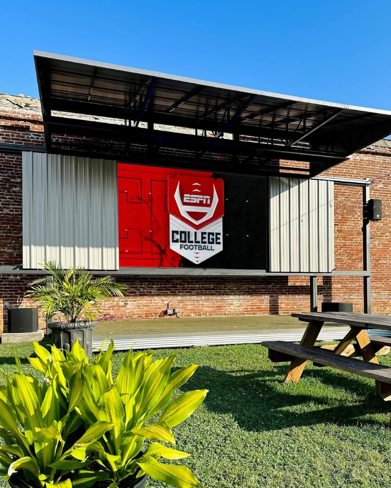 An outdoor screen displays the ESPN College Football logo against a brick wall. A wooden picnic table and green plants are in the foreground, creating a perfect game day ambiance.