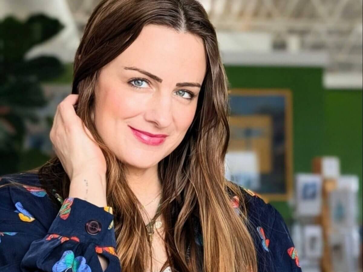 She Brings Art, Vintage Finds & Creativity to Memphis - 2 Woman with long brown hair and a dark floral blouse smiles at the camera, touching her hair, against a blurred green and white indoor background adorned with unique art and vintage finds.