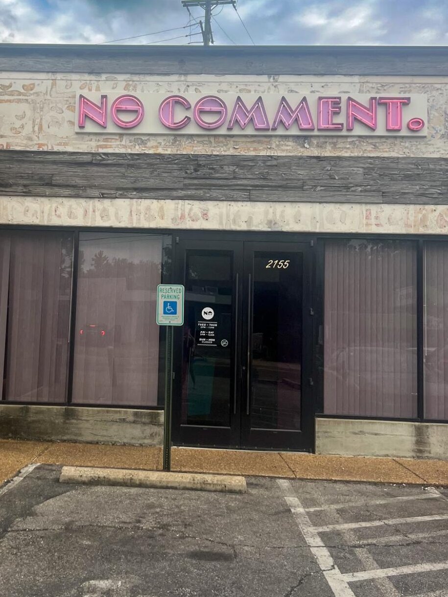 A storefront with a faded exterior sign reading “NO COMMENT.” in pink letters. The sidewalk and parking lot are visible.