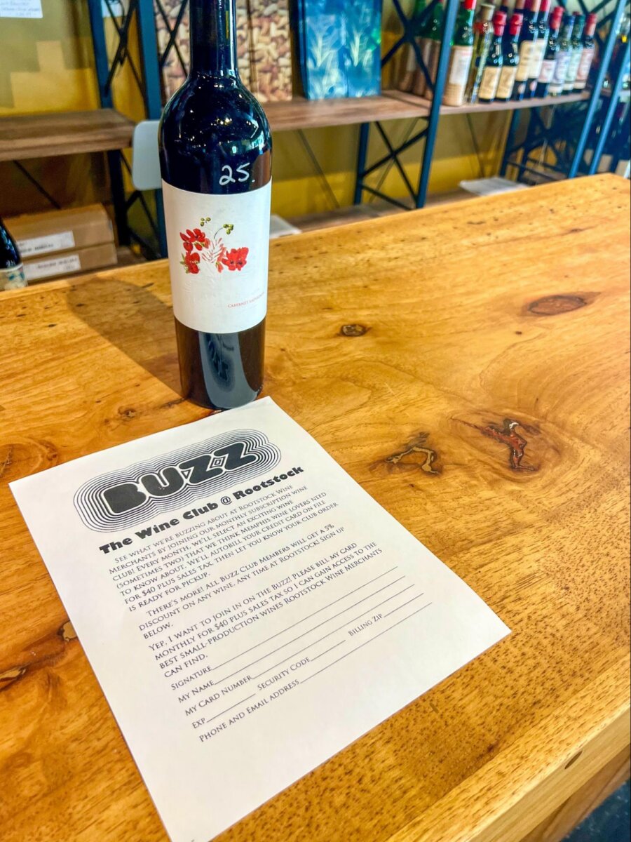 A bottle of red wine labeled “25” sits on a wooden table next to a printed wine club sign-up form at a store with shelves of bottles in the background.