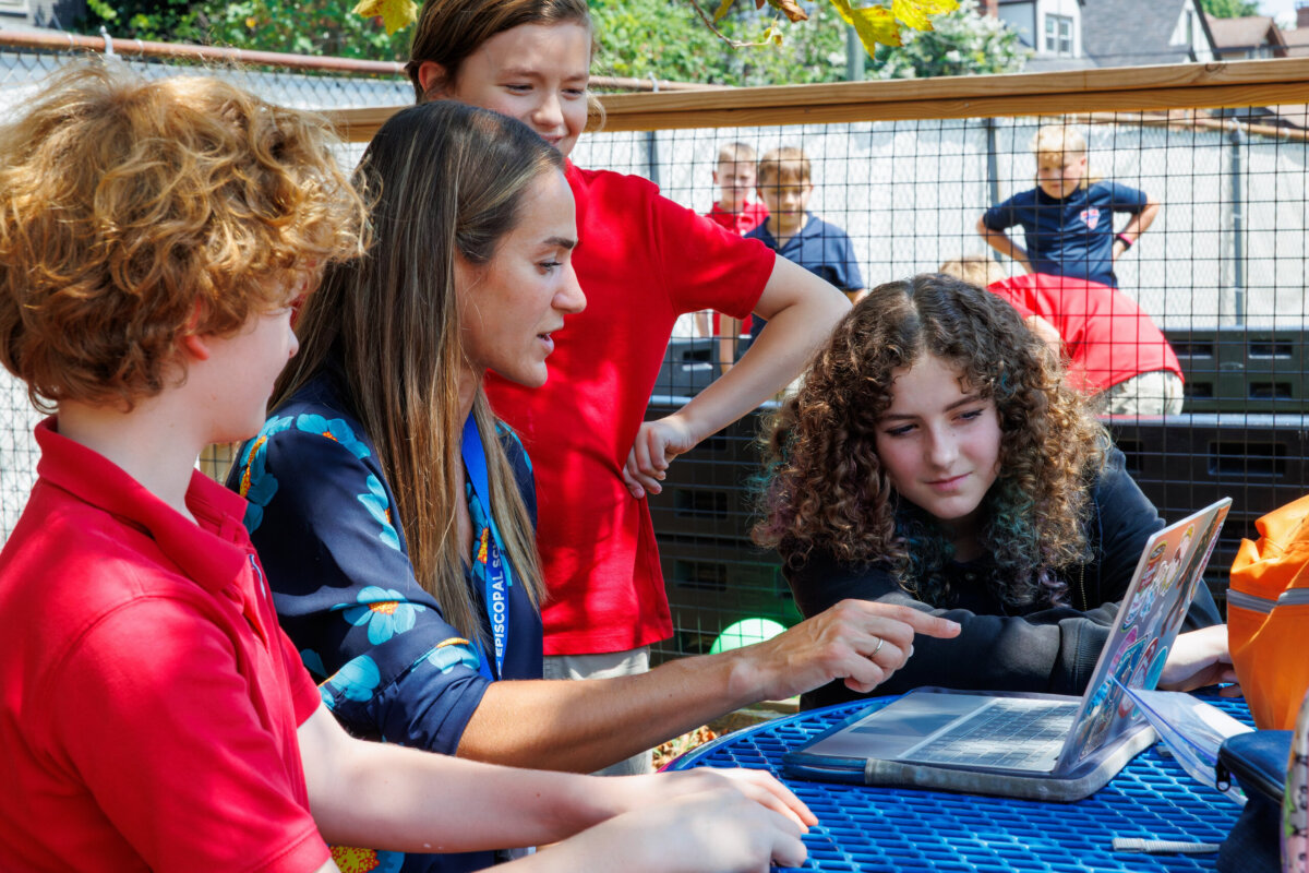 Nashville Private School Open House Dates - 6 A teacher and three students sit at a blue table outside, looking at a laptop screen togetherβa scene that reflects the collaborative learning environment often found in private schoolsβwith more children in the background.