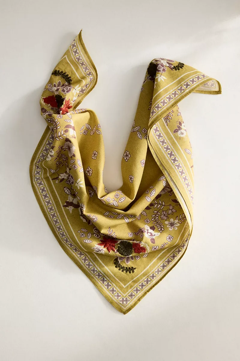 Yellow floral-patterned scarf with a decorative border, arranged in a loose, draped V shape on a light background.