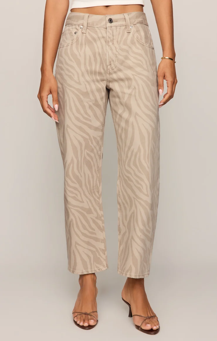 A person wears high-waisted, beige zebra-print jeans with a raw hem, paired with a white crop top, gold bracelet, and heeled sandals.