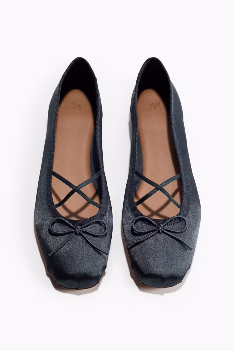 A pair of black satin ballet flats with brown insoles, crisscross elastic straps, and small decorative bows on the front.