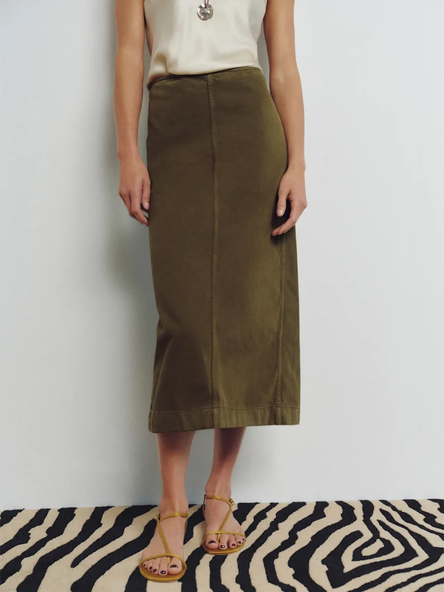 Person wearing an olive green midi skirt, sleeveless beige top, necklace, and yellow sandals, standing on a zebra-print rug against a plain white wall.