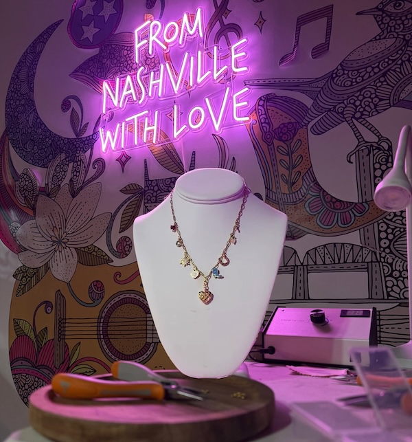 9 New Nashville Retail Spots to Explore - 10 A necklace is displayed on a white stand in front of a wall with a neon sign reading βFrom Nashville With Loveβ and a colorful mural. Jewelry tools and materials are on the table, capturing the creative spirit found in many new retail spots that define the Nashville shopping scene.