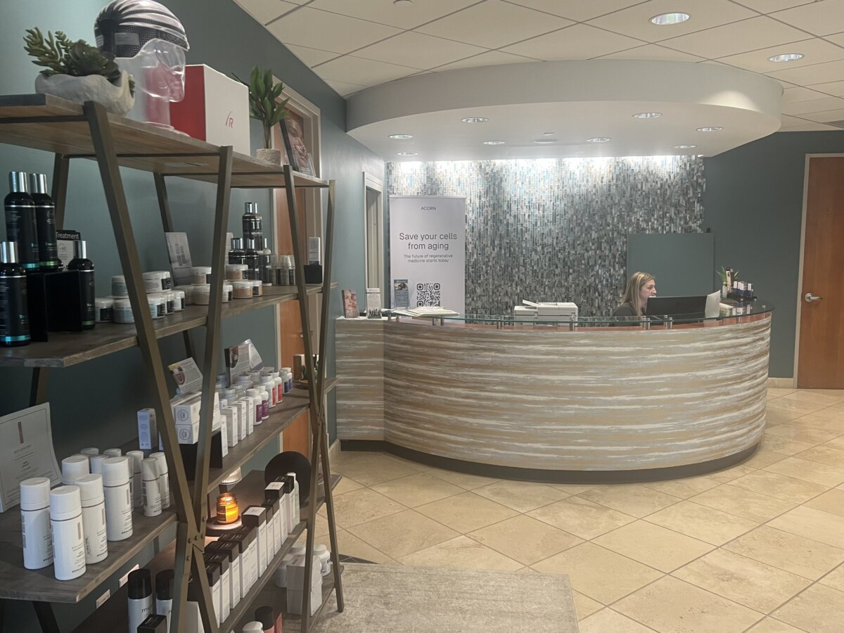 Reception area of a spa or clinic with a curved front desk, a receptionist seated, and shelves displaying skincare products on the left—highlighting summer sales for your favorite treatments and beauty essentials.