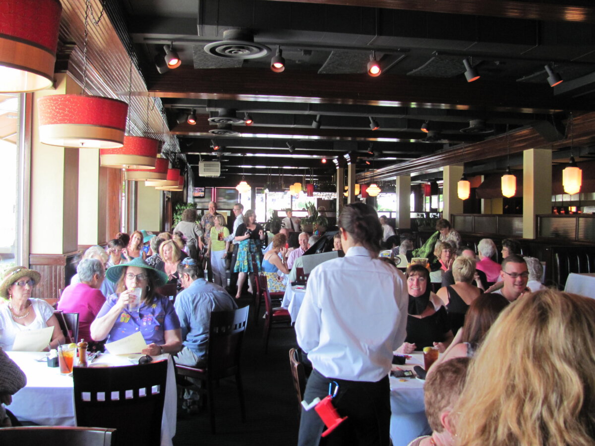 A crowded restaurant dining area with people seated at tables, eating and talking; a server stands in the foreground holding a notepad, while signs throughout the room highlight special fall savings for diners.
