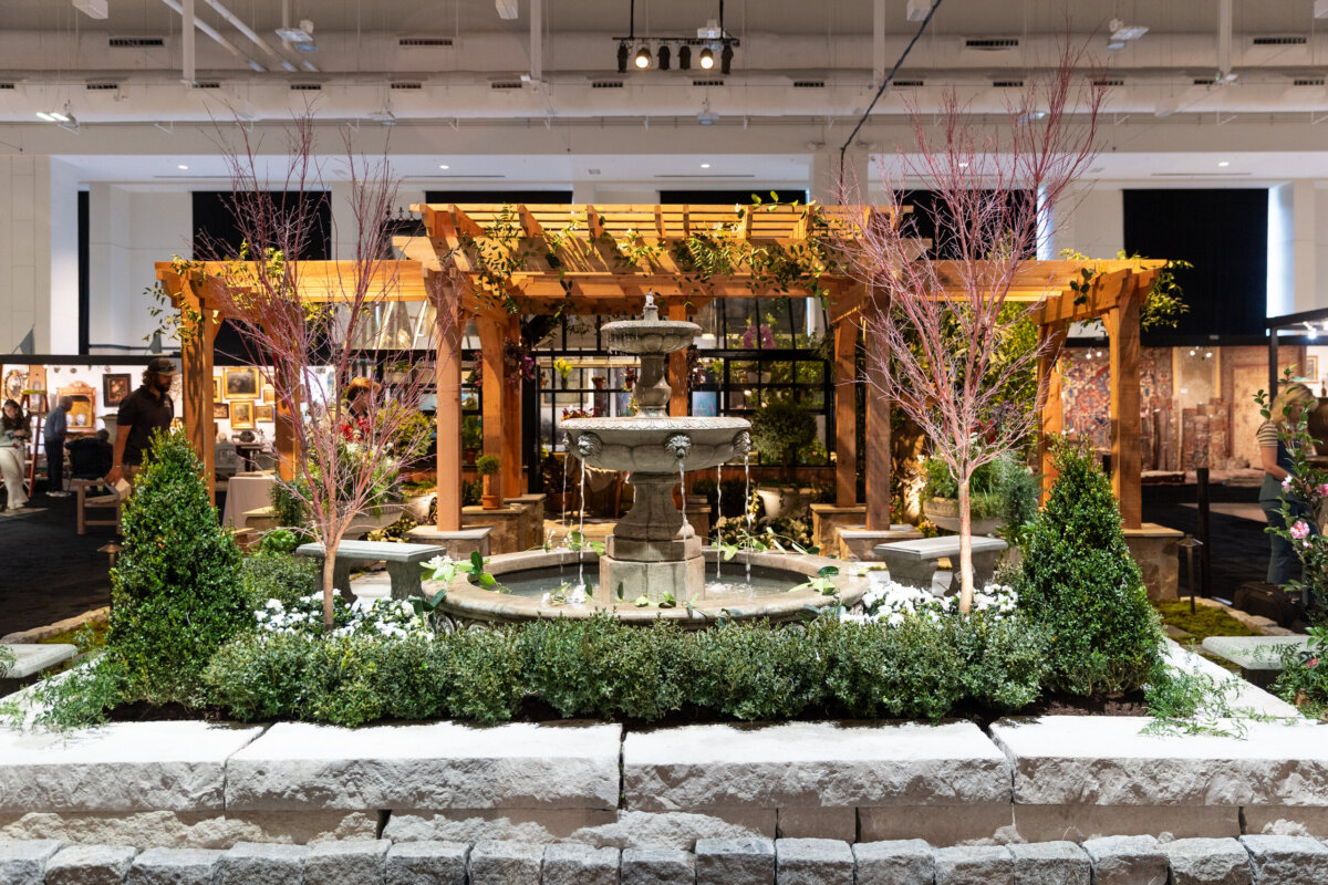 Indoor garden exhibit with a stone fountain, trimmed shrubs, small trees, and a wooden pergola, surrounded by stone edging. Decorative plants and framed art are visible in the background.