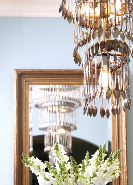 A chandelier made of silverware hangs in front of a large framed mirror, with white flowers in the foreground and a pale blue wall in the background.