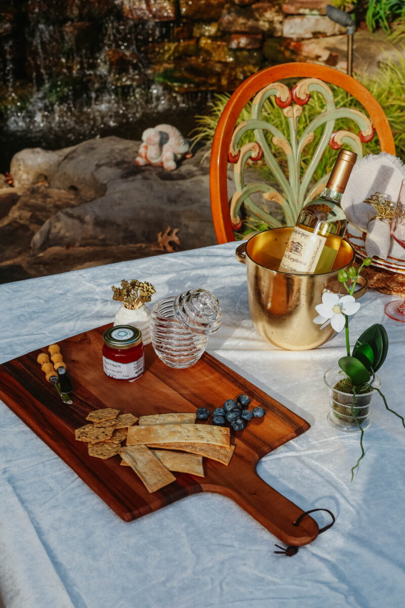 A wooden board with crackers, blueberries, and jam sits on a table next to a gold wine bucket with a bottle—perfect for sharing the latest announcements or celebrating new deals—with a glass vase, a white orchid, and an ornate chair by a rocky waterfall.