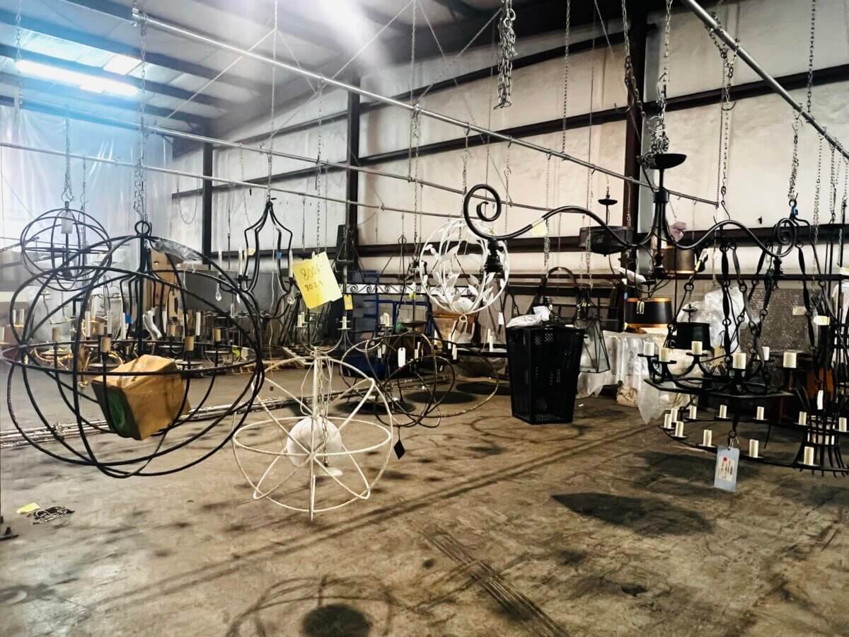 A warehouse interior with several metal chandeliers and light fixtures hanging from the ceiling; some are partially painted, and the space is industrial and organized—perfect for browsing our latest fall savings on lighting solutions.