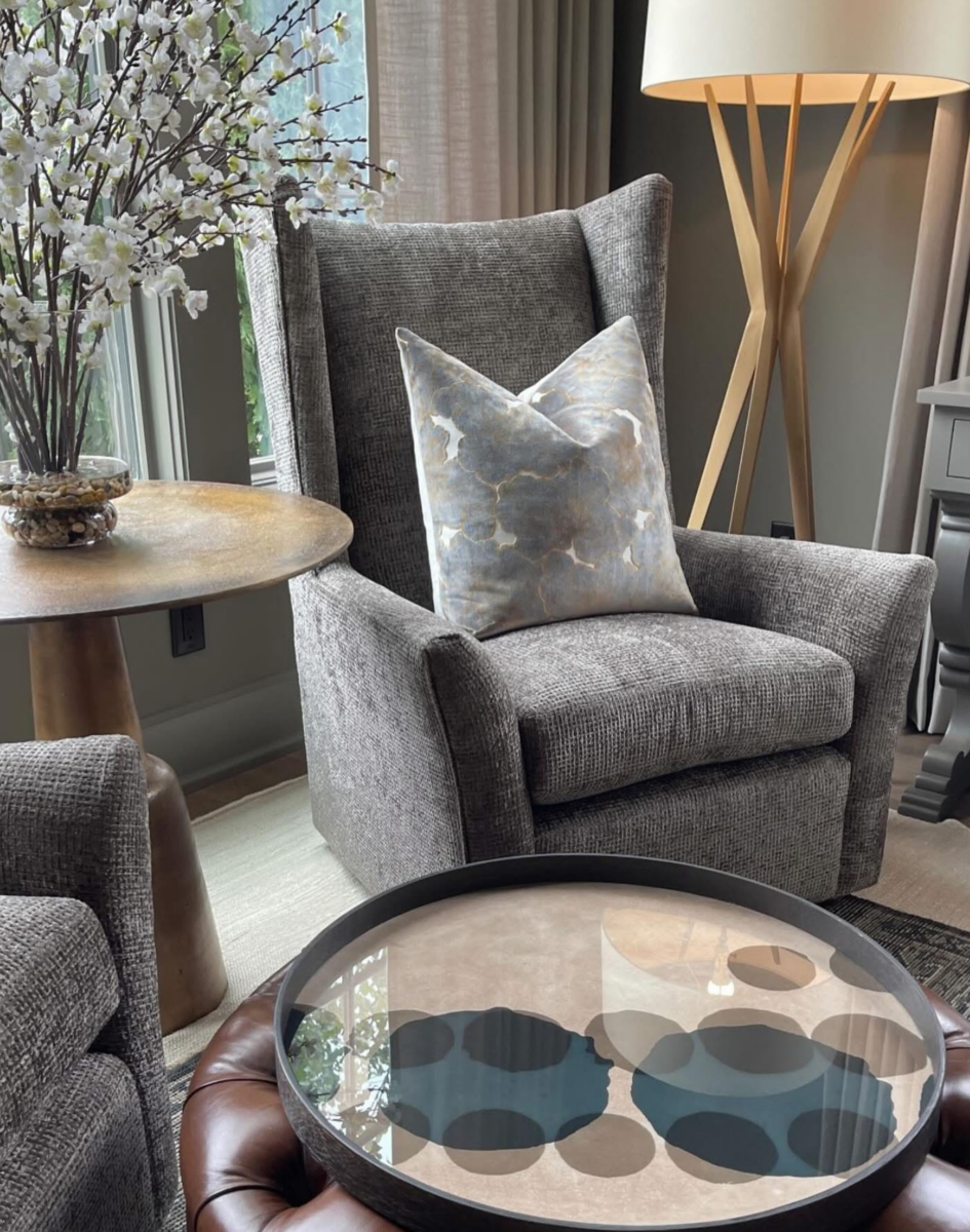 A gray armchair with a patterned pillow sits beside a round wooden table with flowers and a modern floor lamp, near a window in a living room.