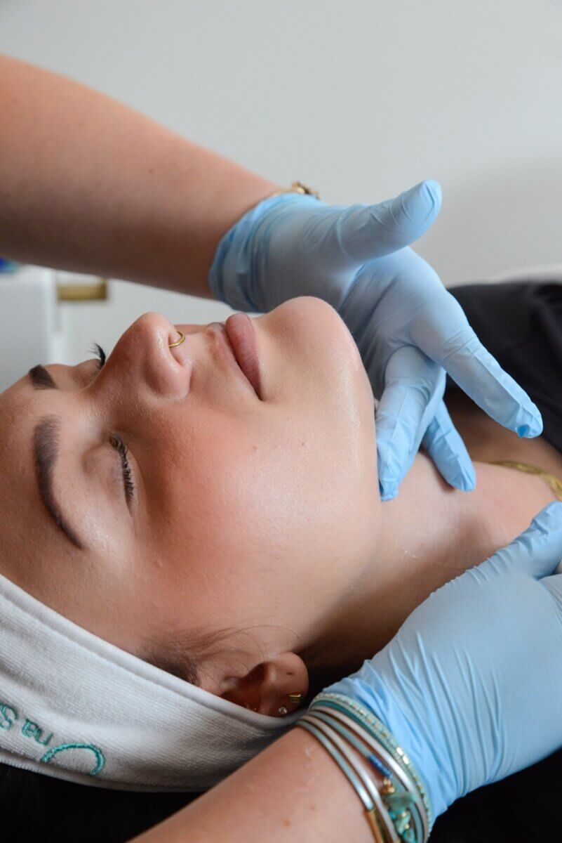 A person with closed eyes enjoys a relaxing facial treatment from someone wearing blue gloves, taking advantage of fall savings. The person has a nose ring and wears a white headband.