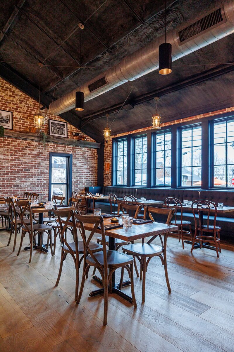 Restaurant dining area with wooden tables and chairs, brick walls, large windows, and industrial-style lighting.